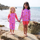 Two children wearing UV Protective Surf Set Pink Starfish, standing on a rock on a beach, smiling at the camera.